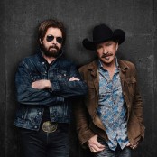 Brooks & Dunn stood against a black wall one wearing a denim jacket and black sunglasses, the other a brown jacket and black cowboy hat